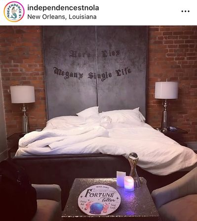 Cozy industrial-chic bedroom with exposed brick, rumpled white bedding, large metal headboard painted with a playful 'single life' message, matching bedside lamps, and a small table at the foot displaying a fortune-teller sign and glowing candles.