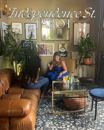 Cozy tarot parlor interior showing a tarot reading: two women seated on a leather sofa and armchair at a glass coffee table with candles and bottles, surrounded by potted plants, framed art and terrazzo floor.