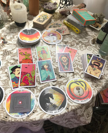 Bright, eclectic tarot and oracle cards spread on a lace-covered table with crystals, a smudge stick, candles and stacked decks — boho home tarot reading setup.