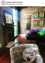 Sunlit New Orleans loft-style room with exposed brick wall and hardwood floors, featuring a black pool table topped with purple and green balloons, a rack of cues and vintage concert posters on the wall, and a striped armchair in the foreground holding a round “Fortune Teller” sign.