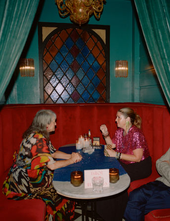 Tarot reading at a red velvet booth in a teal vintage lounge — candles, tarot cards and a stained-glass window behind, reader in a sparkly pink top holding a card over a small round table.