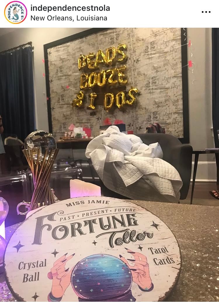 Fortune-teller themed bridal-party setup in New Orleans with a tabletop sign labeled Fortune Teller and crystal-ball graphic, glowing candles and crystal ball prop in foreground, cozy seating, and gold 'Beads Booze & I Dos' balloon letters on the wall.