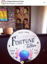 Quirky round “Miss Jamie Fortune Teller” sign with illustrated crystal ball and hands reading “Crystal Ball” and “Tarot Cards,” perched on a pool table in a New Orleans bar interior with exposed brick, a vintage poster and pop‑art portraits on the wall.