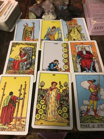 Bright illustrated tarot card spread showing pentacles, swords and wands laid out on a wooden table with healing crystals and a card box for a tarot reading.
