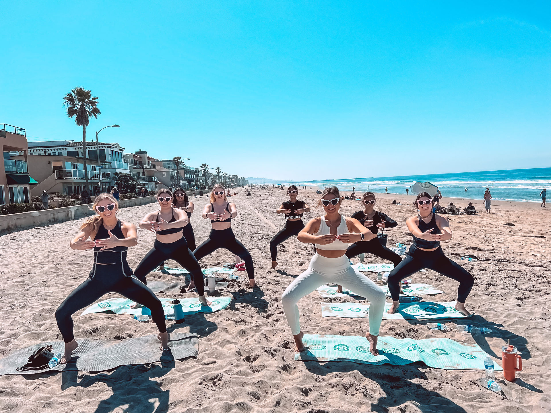 Group beach workout: women in athletic wear and heart-shaped sunglasses doing wide-legged squats on yoga mats at a sunny coastal California beach with ocean waves and beachfront homes in the background.