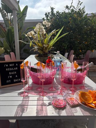 Sunlit backyard patio bachelorette brunch setup with pink plastic champagne flutes garnished with orange peels, two pink ice buckets with bottles on ice, a sign reading "I'M NOT A REGULAR BACH I'M A FIT BACH", orchid centerpiece, sliced oranges and cookies, tropical plants and pink chairs in the background.