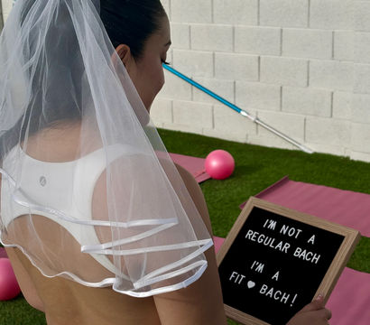 Woman in a bridal veil and white workout wear holding a letterboard that reads "I'M NOT A REGULAR BACH I'M A FIT ♥ BACH!" at a backyard bachelorette fitness setup with pink yoga mats, a small pink exercise ball, and a pool skimmer against a cinder block wall.