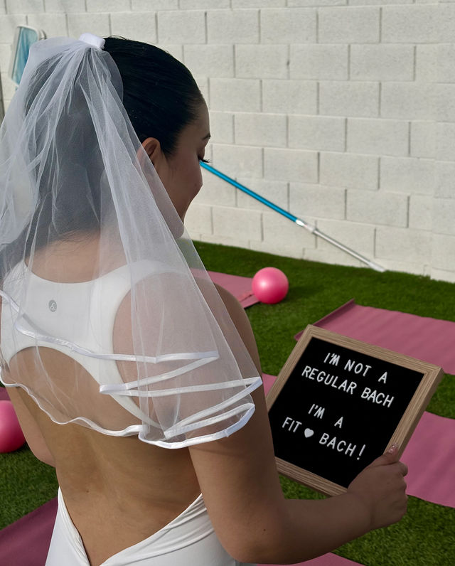 Woman in a bridal veil and white workout wear holding a letterboard that reads "I'M NOT A REGULAR BACH I'M A FIT ♥ BACH!" at a backyard bachelorette fitness setup with pink yoga mats, a small pink exercise ball, and a pool skimmer against a cinder block wall.