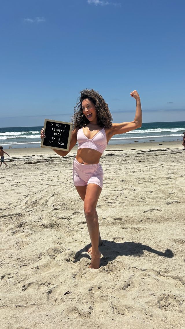 Smiling woman in a pink workout set flexing her arm and holding a letterboard that reads “Not a regular beach, I’m a fit beach” on a sunny sandy beach with ocean waves in the background.