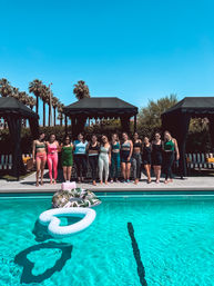 Group of women in colorful activewear lined up poolside in front of black cabanas and palm trees under a clear blue sky, turquoise pool with heart-shaped and tropical-print inflatables