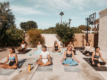 Sunny outdoor yoga class with women seated in meditation on mats on a suburban patio, palm trees and a basketball hoop in the background