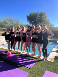 Eight women in activewear posing barefoot poolside on a sunny day for a backyard outdoor workout — purple yoga mats and a pink exercise ball on the turf, one holding a small letterboard.