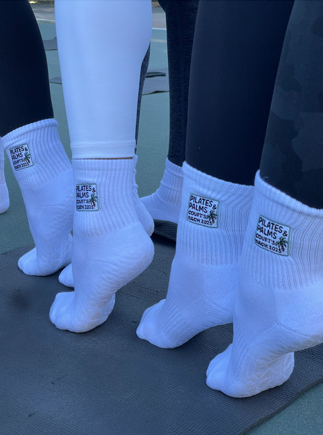 Close-up of several people on an outdoor court standing on a yoga mat wearing white grippy ankle socks with embroidered patches and leggings, giving a group pilates/yoga class vibe.