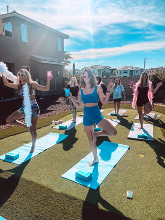Outdoor backyard yoga party in a sunny suburban neighborhood: group of women in colorful activewear and heart-shaped sunglasses striking tree pose on blue mats on artificial turf, holding cups and wearing feather boas.