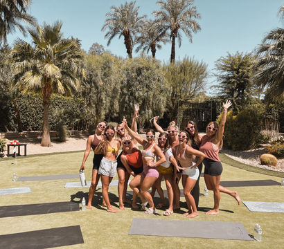 Group of women in sunglasses posing and smiling on yoga mats on a sunny lawn surrounded by palm trees and desert landscaping - outdoor yoga/retreat vibe.