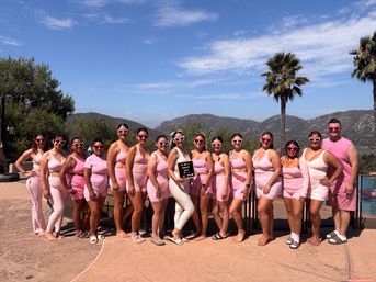 Cheerful group wearing matching pink outfits and heart-shaped sunglasses posing on a sunny poolside deck with palm trees and rolling hills in the background, one person holding a small sign.
