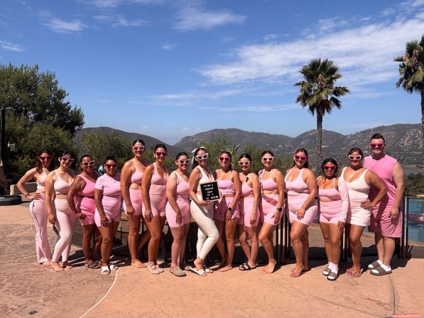 Cheerful group wearing matching pink outfits and heart-shaped sunglasses posing on a sunny poolside deck with palm trees and rolling hills in the background, one person holding a small sign.