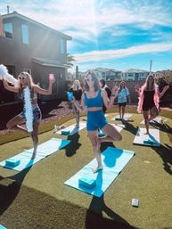 Outdoor backyard yoga party in a sunny suburban neighborhood: women in colorful activewear and heart-shaped sunglasses balance in tree pose on blue mats, holding drinks and feather boas.