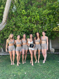 Six women barefoot on grass in a sunny backyard, smiling in matching neutral sports bras and bike shorts (center wearing white) standing in front of a dense green hedge, holding a small letterboard sign — casual group fitness/garden photo.