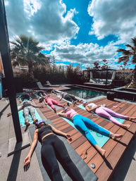 Relaxing outdoor poolside yoga session with a group lying on mats on a wooden deck, palm trees and lounge chairs beside a pool under a bright blue sky with dramatic clouds.