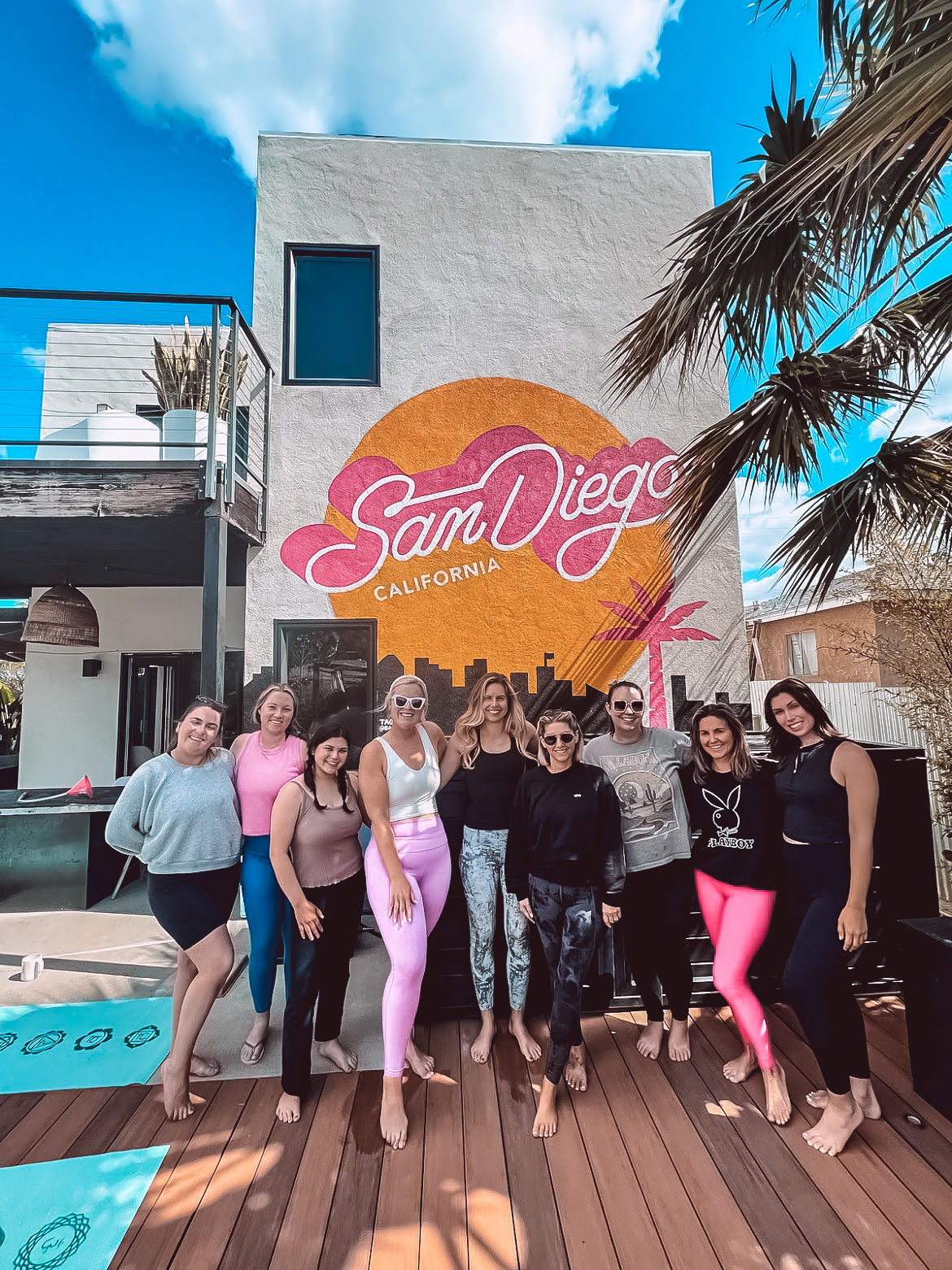 Group of women posing barefoot on a sunny San Diego wooden deck in front of a colorful "San Diego, California" mural, with palm trees and yoga mats nearby.