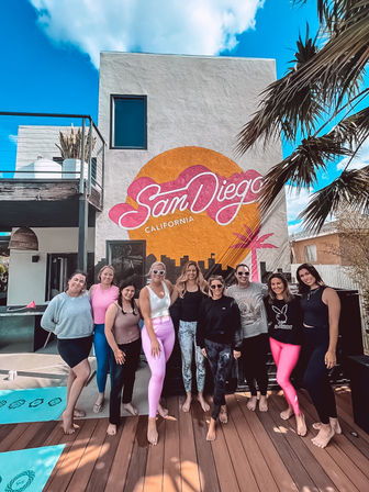 Group of women posing barefoot on a sunny San Diego wooden deck in front of a colorful "San Diego, California" mural, with palm trees and yoga mats nearby.
