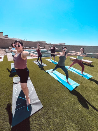 Group of women wearing heart-shaped sunglasses practicing Warrior II yoga on mats on a sunny backyard lawn next to a pool under a clear blue sky.