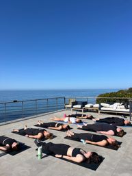 Oceanfront terrace yoga: group lying in Savasana on mats on a sunlit coastal patio with clear blue sky and calm sea horizon