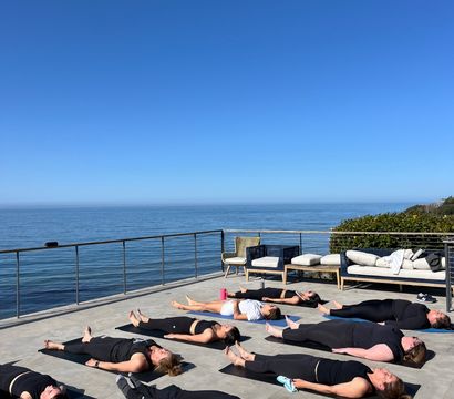 Oceanfront terrace yoga: group lying in Savasana on mats on a sunlit coastal patio with clear blue sky and calm sea horizon