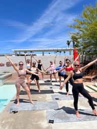 Outdoor poolside yoga class in Palm Springs: women in activewear striking energetic poses on patterned mats beside a turquoise pool under a bright blue sky with palm trees.