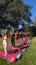 Outdoor fitness group on a sunny backyard lawn: women in activewear and heart-shaped sunglasses lined on pink yoga mats with pink balloons and rolled towels beneath a large oak tree.