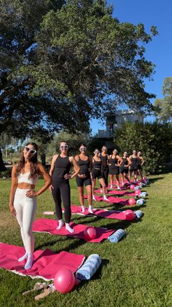 Outdoor fitness group on a sunny backyard lawn: women in activewear and heart-shaped sunglasses lined on pink yoga mats with pink balloons and rolled towels beneath a large oak tree.