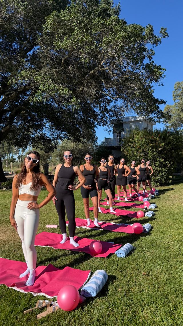 Outdoor fitness group on a sunny backyard lawn: women in activewear and heart-shaped sunglasses lined on pink yoga mats with pink balloons and rolled towels beneath a large oak tree.