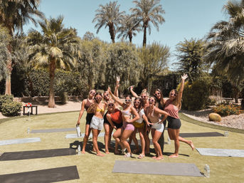 Group of women in sunglasses posing and smiling on yoga mats on a sunny lawn surrounded by palm trees and desert landscaping - outdoor yoga/retreat vibe.