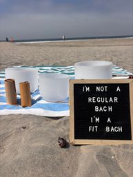Frosted crystal singing bowls and wooden mallets on a blue-and-white striped towel at a sandy ocean beach, beside a letter board reading 'I'm not a regular Bach I'm a fit Bach'.