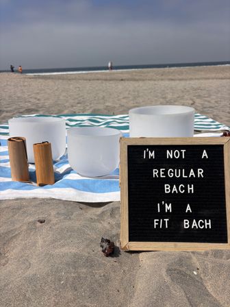 Frosted crystal singing bowls and wooden mallets on a blue-and-white striped towel at a sandy ocean beach, beside a letter board reading 'I'm not a regular Bach I'm a fit Bach'.
