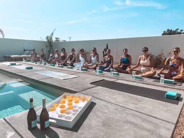 Sunny poolside yoga session with a line of women in activewear seated in meditation on mats along a modern concrete deck, tray of orange cocktails and two bottles by the pool, relaxed outdoor wellness vibe.
