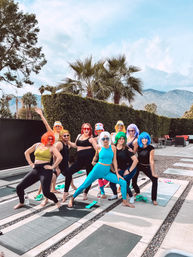 Outdoor yoga group of adults in colorful wigs and activewear striking playful poses on mats at a poolside patio, framed by palm trees and a mountain backdrop.