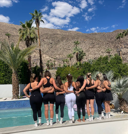 Eight women in workout clothes standing arm‑in‑arm on a pool deck beside a turquoise pool, facing a rocky desert mountain and tall palm trees under a bright blue sky — lively desert oasis group shot.
