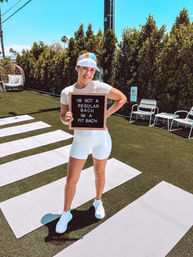 Smiling woman in white workout gear and sun visor on a sunny outdoor lawn with yoga mats and palm trees, holding a letterboard that reads “IM NOT A REGULAR BACH IM A FIT BACH”