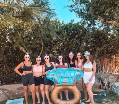 Seven women in sporty summer outfits and colorful visors pose around a giant inflatable diamond ring in a sunny backyard with palm trees, green hedge, yoga mats and water bottles.