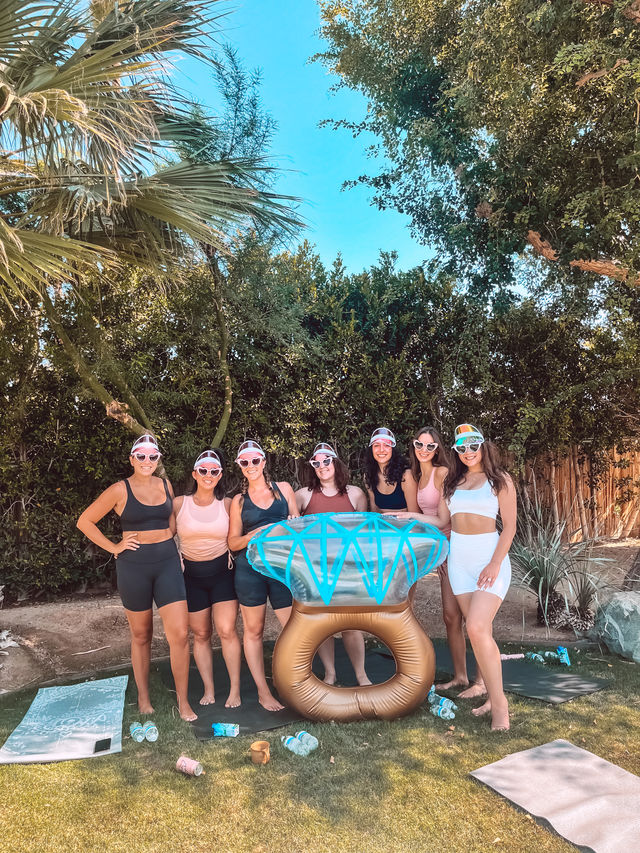 Seven women in sporty summer outfits and colorful visors pose around a giant inflatable diamond ring in a sunny backyard with palm trees, green hedge, yoga mats and water bottles.
