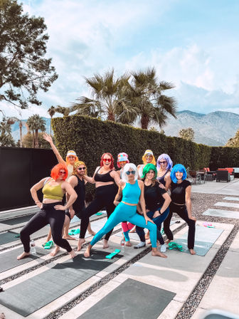 Outdoor yoga group of adults in colorful wigs and activewear striking playful poses on mats at a poolside patio, framed by palm trees and a mountain backdrop.