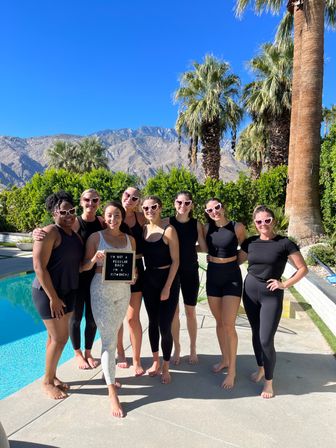 Nine women in matching activewear and heart-shaped sunglasses pose barefoot by a pool with palm trees and desert mountains in the background; one holds a letterboard that reads I'm not a regular bach I'm a fit bach.