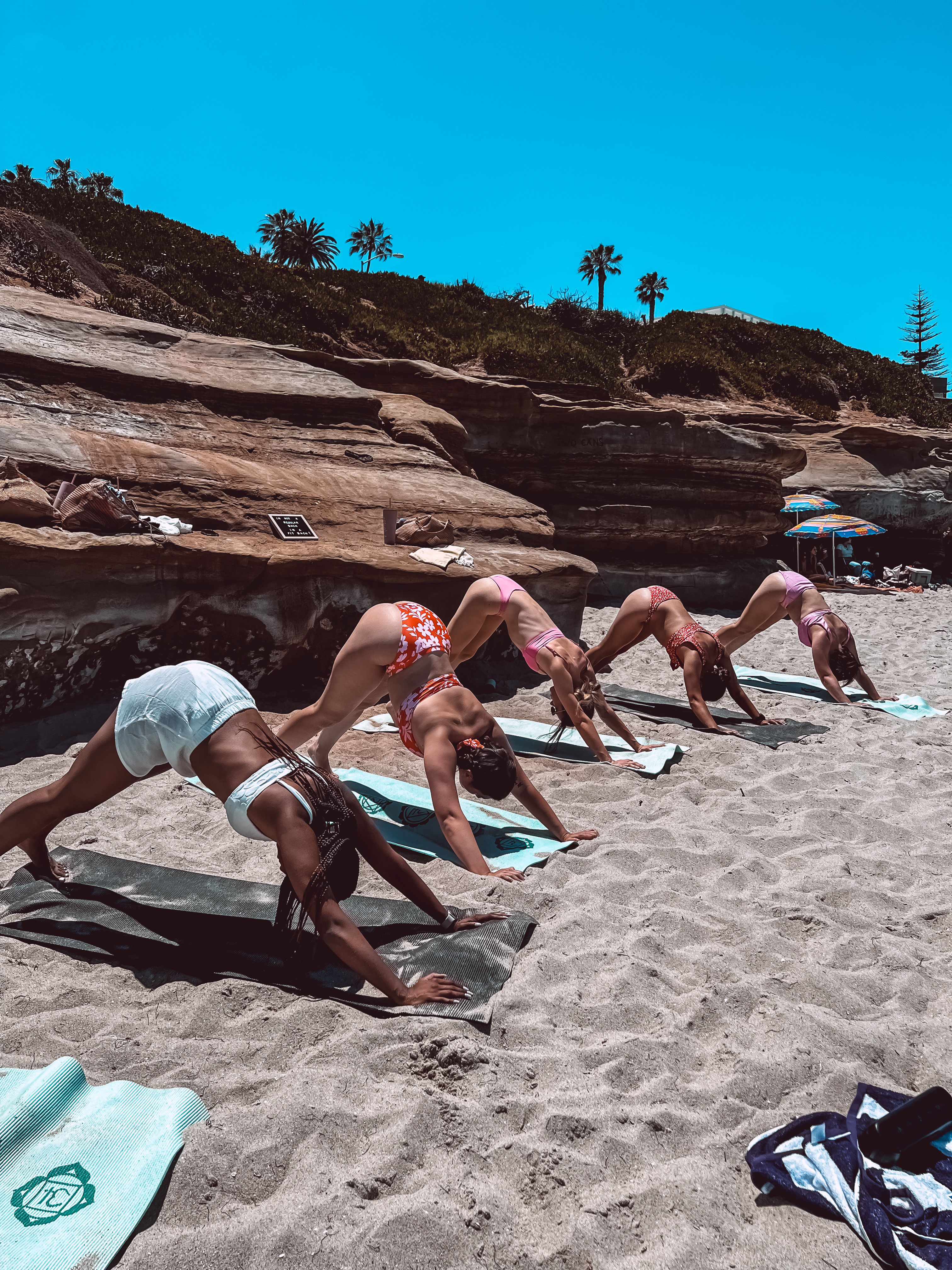 Five women in swimsuits practicing downward dog on yoga mats on a sunny sandy beach by sandstone cliffs and palm trees — outdoor seaside yoga session.