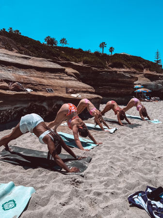 Five women in swimsuits practicing downward dog on yoga mats on a sunny sandy beach by sandstone cliffs and palm trees — outdoor seaside yoga session.