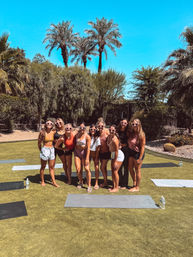 Group of women wearing heart-shaped sunglasses posing on yoga mats on a sunny, palm‑lined lawn with desert landscaping — outdoor yoga gathering in a backyard garden.
