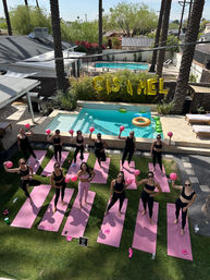 Poolside outdoor yoga class of women on pink mats holding pink exercise balls beneath palm trees beside a backyard pool with gold letter balloons.
