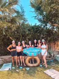 Seven women in sporty summer outfits and colorful visors pose around a giant inflatable diamond ring in a sunny backyard with palm trees, green hedge, yoga mats and water bottles.