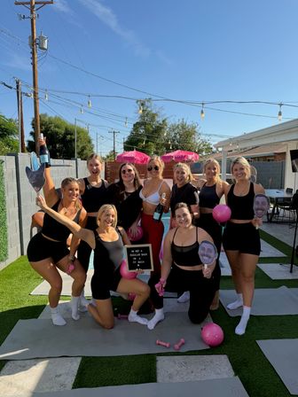 Ten women in black workout outfits posing by a sunny backyard pool on a residential patio, holding pink exercise balls, small dumbbells, a champagne bottle, face cutouts and a letterboard — playful outdoor fitness-style pool party.